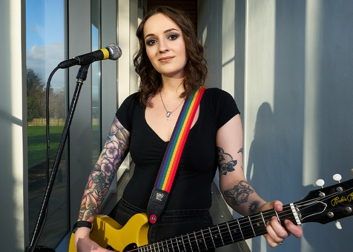 A young woman stands in front of a microphone stand, poised to play an electric guitar, in a corner of a glass fronted recording studio